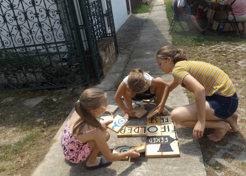 Stifolder fesztivál - a "Hagyományok udvara" előtt gyerekek óriás fa kirakóval játszanak, a 20 darabos puzzle egyik oldala Feked címerét, másik oldala a Stifolder fesztivál logóját ábrázolja